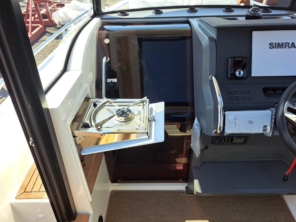 Interior of a boat showing a mounted SIMRAD display, a stove, and control panel with buttons and a fabric-wrapped steering wheel.